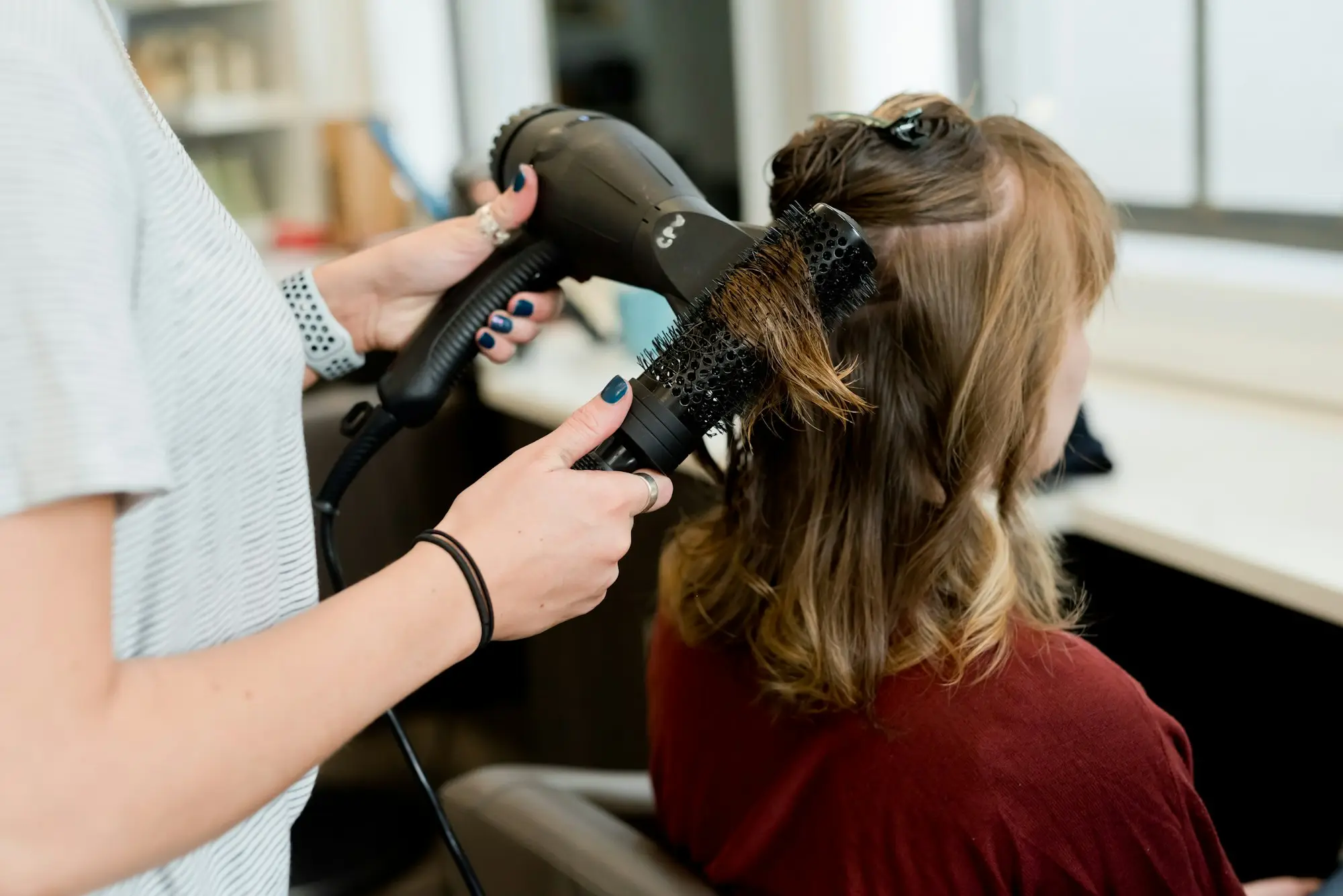 Hairdresser blow-drying a client's hair with a round brush at her station in a small salon