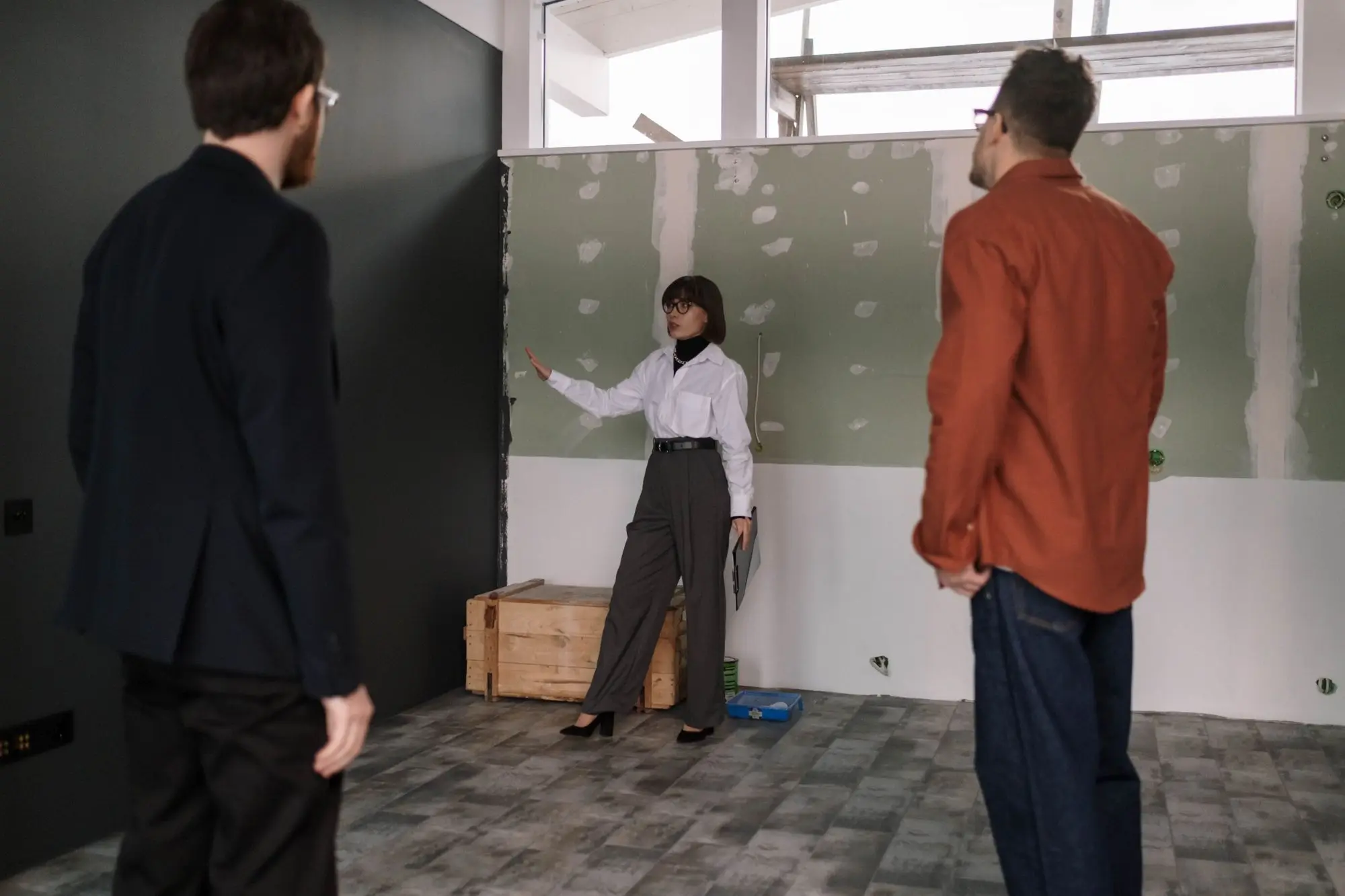 Property manager walking two prospective tenants through a shop tenancy mid fit-out, with patched plasterboard walls and a timber crate on the floor